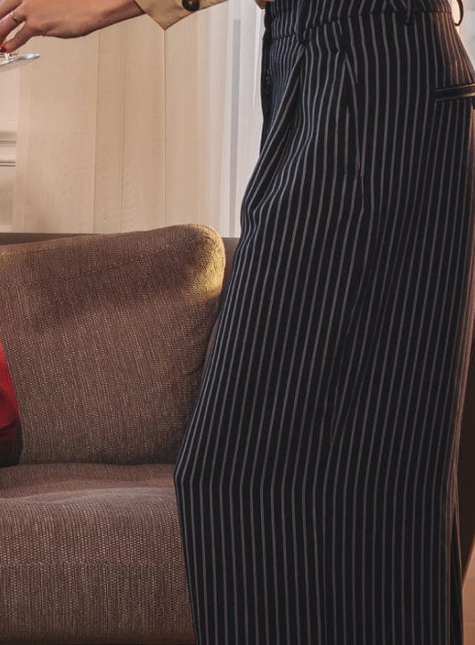 A photo of a woman in black trousers with white stripes holding a glass, brown sofa in the background