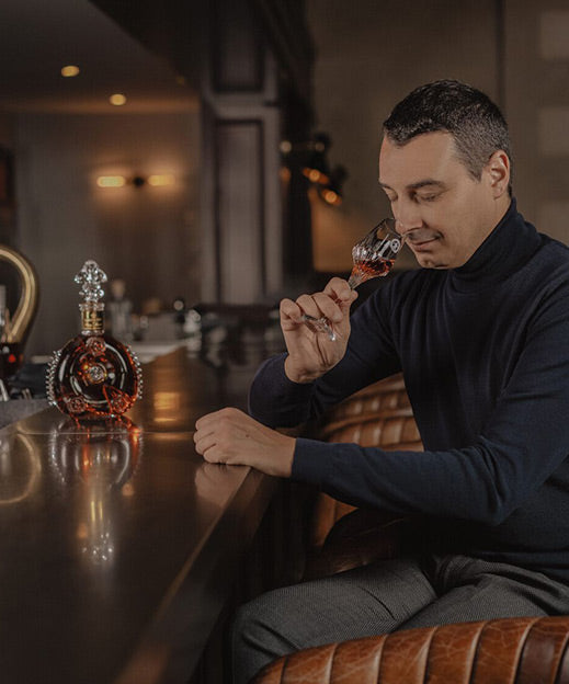 A lifestyle photo of Baptiste Loiseau smelling LOUIS XIII from a crystal glass, decanter on the bar countertop
