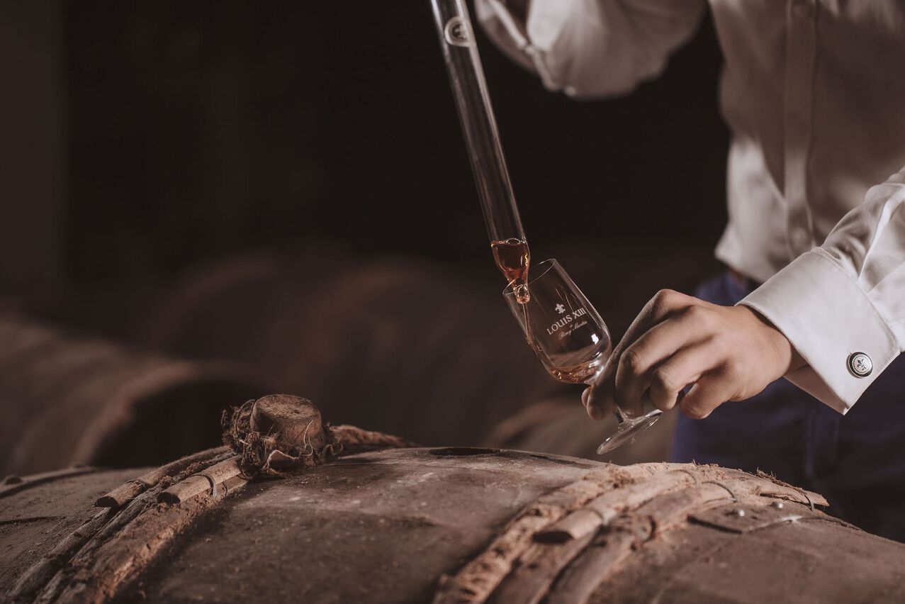 A lifestyle photo of a hand holding a glass and filling it with cognac using a spear, a barrel below