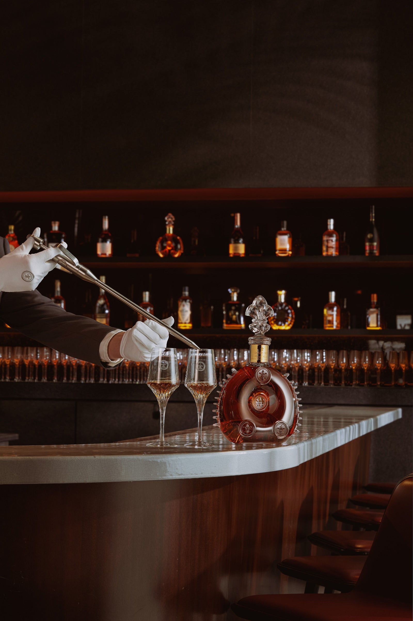 A lifestyle photo of a bartender serving LOUIS XIII to two crystal glasses with a spear, decanter nearby