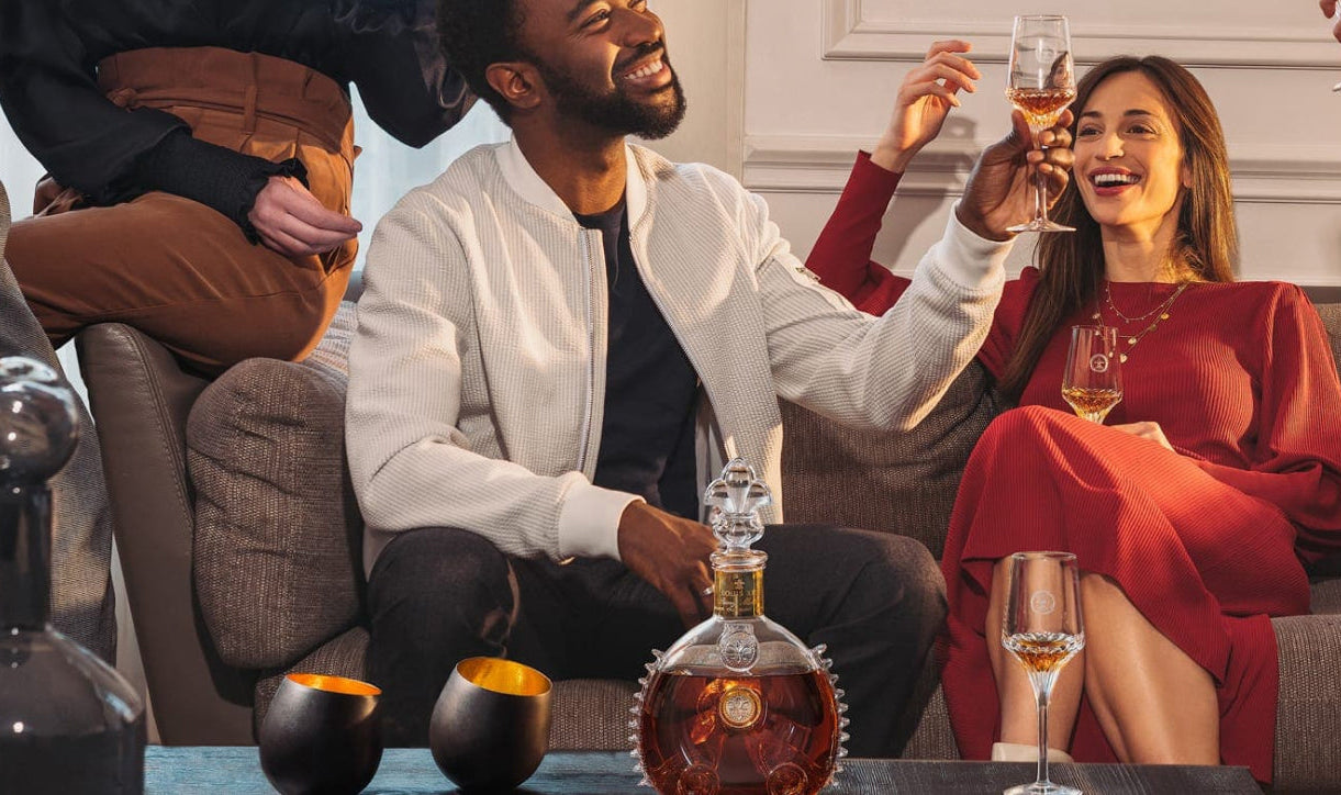 A lifestyle photo of two people on a sofa toasting with LOUIS XIII crystal glases, a decanter and a glass on the table