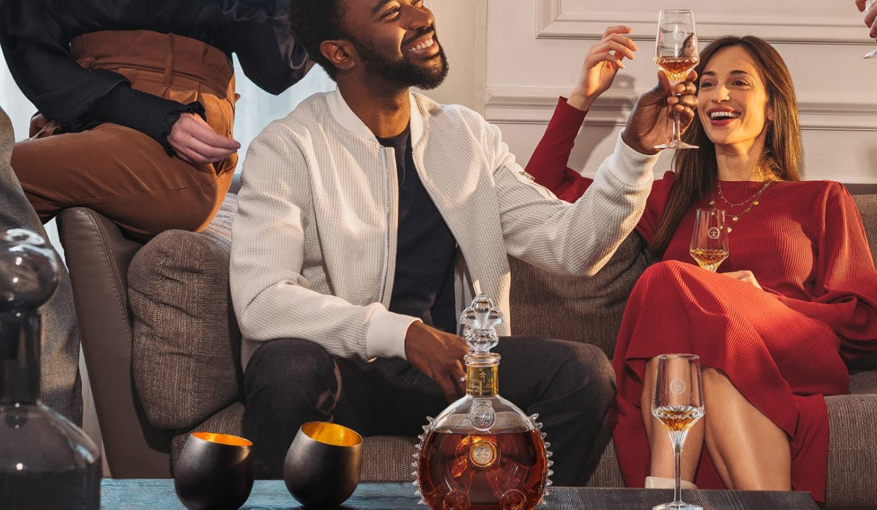 A lifestyle photo of two people on a sofa toasting with LOUIS XIII crystal glases, a decanter and a glass on the table