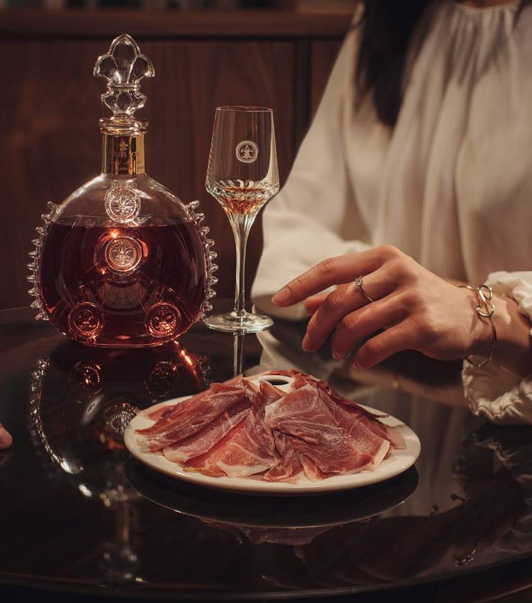 A lifestyle photo of a hand reaching bellota ham, a decanter and crystal glass in the background