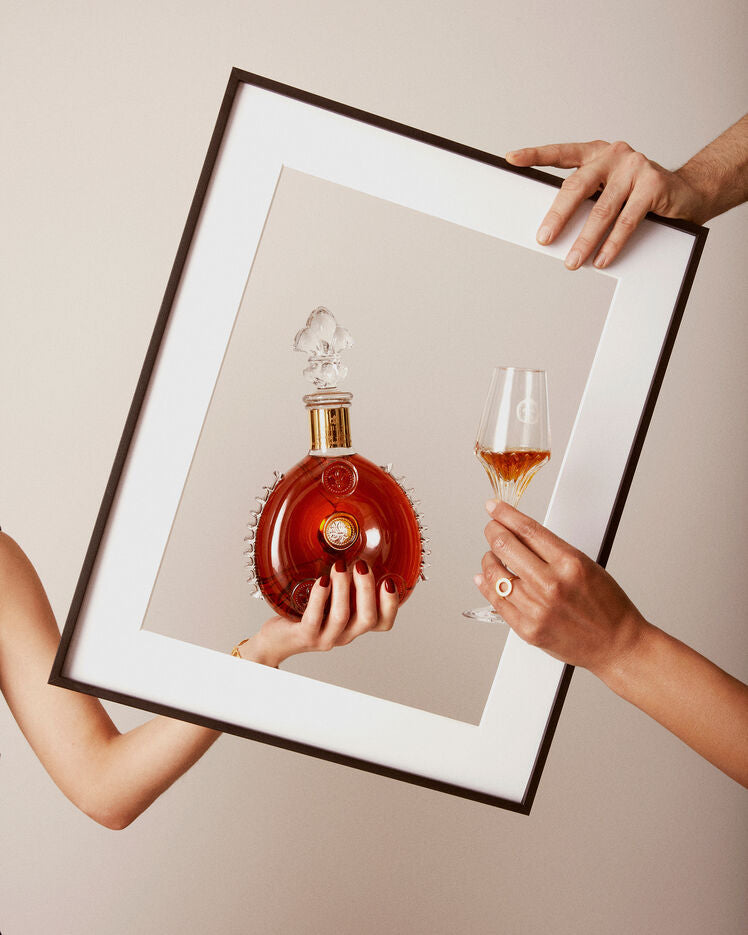 A lifestyle image of a black frame, behing a hand holding LOUIS XIII decanter and other hand holding LOUIS XIII crystal glass