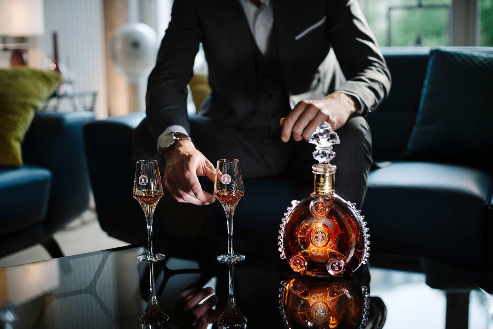 A lifestyle image of a man reaching for LOUIS XIII crystal glass, another glass and a decanter on the table