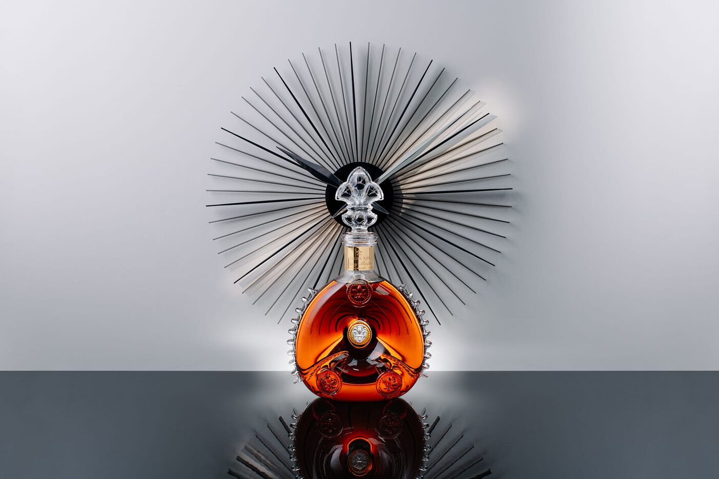 A lifestyle photo of LOUIS XIII decanter on a black shiny surface, a black modern clock behind