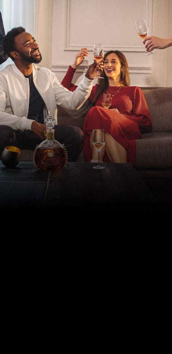 A lifestyle photo of two people on a sofa toasting with LOUIS XIII crystal glases, a decanter and a glass on the table