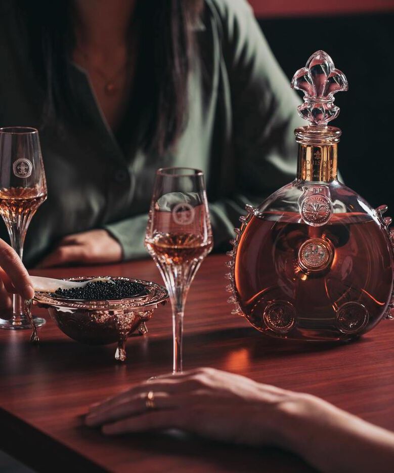 A lifestyle photo of a female hand holding LOUIS XIII crystal glass, a bowl of black caviar and a decanter in the background