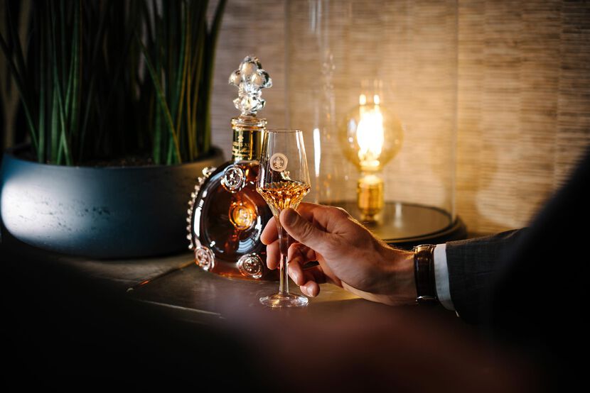 A lifestyle photo of a hand holding LOUIS XIII crystal glass, decanter behind, a lampwith yellow light and a bamboo plant in the background
